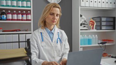 Hasta bakımı çözümleri hakkında araştırma ve veri girişi için dizüstü bilgisayar kullanırken tıbbi ekipman ve dosyalarla klinikte çalışan kadın doktor.