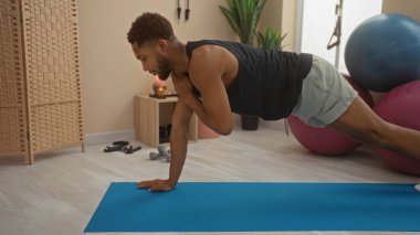 Spor salonunun arka planında spor malzemeleriyle mavi bir paspasın üzerinde egzersiz yapan genç bir adam. Forma, kuvvete ve dengeye odaklanıyor.