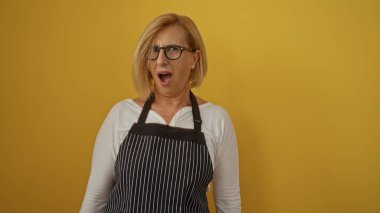 Sarı arka planda duran, şaşırmış bir ifadeyle önlüklü, olgun sarışın kadın.