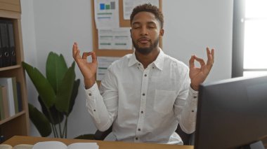 Genç adam iş yerinde meditasyon yaparken, gözleri kapalı, avuçları havada, beyaz gömlek giymiş, dizüstü bilgisayar, kâğıt ve bitki gibi arka planda.