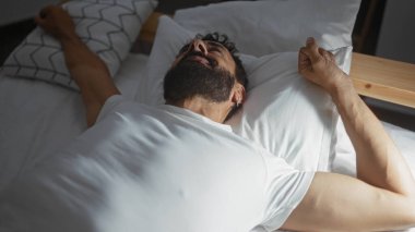 Genç İspanyol bir adam rahat bir yatak odasında uzanmış, sabah uyandığında kollarını uzatıyor.