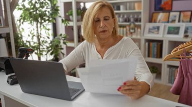 Yetişkin bir kadın, arka planda raflar ve bitkiler olan bir ev dekoratörü masasında belgeleri analiz ediyor.