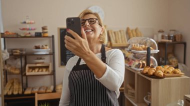 Kadın, çeşitli hamur işleri ve ekmeklerle dolu fırında selfie çekerken gülümsüyor. Etrafı canlı fırın dekorlarıyla çevrili, fırında pişmiş yiyecekler ve rahat bir atmosfer sergiliyor..