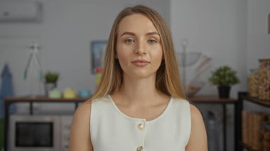 Ofiste, modern dekorla ayakta duran bir kadın, kendine güvenen, sarı saçlı, sıradan giyinen ve tarafsız bir yüz ifadesi olan genç bir kadın..