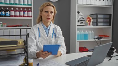 Klinikteki kadın doktor beyaz önlük giyip raflarda tıbbi ekipmanlarla tablet kullanıyor. Bu da hastane ortamında çalışan bir doktor olduğunu gösteriyor..