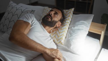 Yakışıklı, sakallı, genç bir adam. Parlak yatak odasında, iç tarafta, desenli yastıklarla yatıyor..