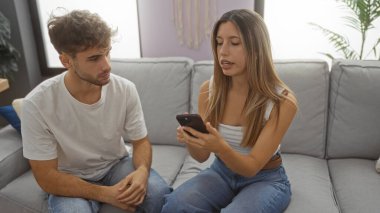 Modern bir oturma odasındaki kanepede oturan kadın ve erkek ellerinde akıllı bir telefonla sohbet ediyorlar. Kapalı alan ortamını resmediyorlar..