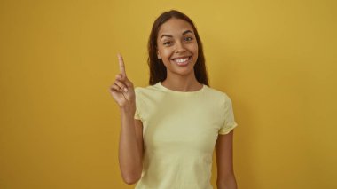 Güzel, genç İspanyol bir kadın gülümsüyor ve sarı bir duvara işaret ediyor. Neşeli bir havayla izole edilmiş bir arka plan.