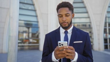 Takım elbiseli genç adam, arka planda modern mimari binalar olan bir şehir caddesinde dikilirken akıllı telefon kullanıyor.