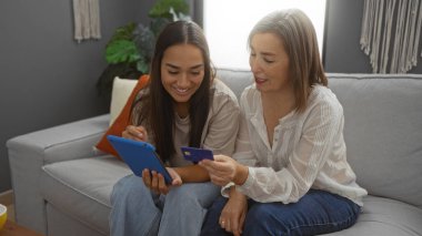 Anne ve kızı oturma odasında birlikte gülümsüyor, tablet ve kredi kartı kullanıyor, aile sevgisini, dostluğu ve modern ev ortamını kapalı alanlarda kadınlara gösteriyor.