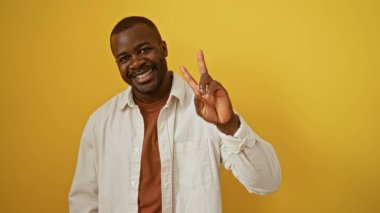 Genç, yakışıklı, Afrikalı, Amerikalı adam gülümsüyor ve izole edilmiş sarı arka planda barış işareti yapıyor..