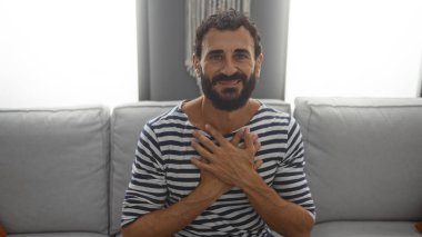 Sakallı, çizgili tişörtlü ve rahat bir oturma odasındaki kanepede oturan yakışıklı bir genç.