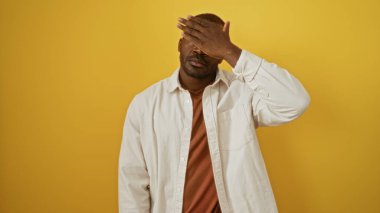 Tek eliyle yüzünü kapatan genç bir adam izole edilmiş sarı arka planda.