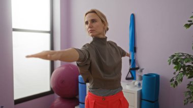 Spor salonunda yoga yapan bir kadın pembe egzersiz topları ve mavi paspaslarla mor renkli bir arka planda egzersiz yapıyor.