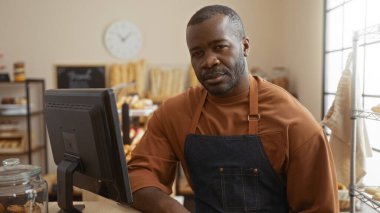 Genç Afro-Amerikalı bir adam pastanede çalışıyor. Kapalı mekanda önlük takıyor, içini ekmek rafları ve poz sistemi ile donatıyor..