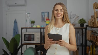 Modern ofiste elinde tabletle gülümseyen bir kadın, bitki, mutfak gereçleri ve raflarla profesyonel çevreyi sergiliyor, genç yetişkin beyaz görünümünü vurguluyor..