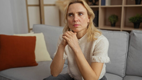 Woman sitting on a modern sofa in a cozy living room, deep in thought with a calm expression, surrounded by soft cushions and ambient indoor lighting.