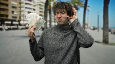 Şehirde İspanyol asıllı bir adam, şehir binaları ve palmiye ağaçları tarafından çevrelenmiş bir jest yaparken Czech Koruna banknotlarını elinde tutuyor..