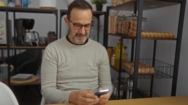 Modern mutfakta akıllı telefon kullanan olgun bir İspanyol adam rafları, mikrodalgası ve kahve makinesi olan, ekrana odaklanmış, gözlük ve süveter giyen, rahat bir atmosferi olan..