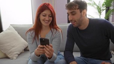 Kadın ve erkek kanepede akıllı telefon kullanarak evde eğleniyorlar, rahat bir oturma odasında mutlu ilişkilerini vurguluyorlar..