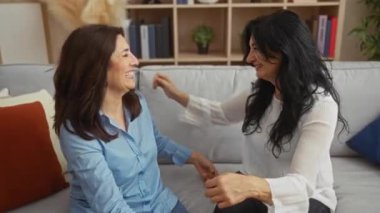 İki yetişkin kadın neşeli bir şekilde rahat bir salonda rahat bir koltukta kucaklaşıyorlar. Aile sevgisini ve dostluğunu içeride mutluluk ve rahatlıkla ifade ediyorlar..