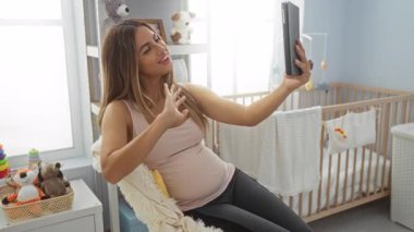 Hamile kadın yatak odasında beşik ve pelüş hayvanlarla sohbet ediyor, evde huzurlu bir ortam sergiliyor..