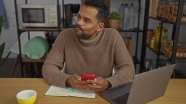 Ofis ortamında telefonu kullanan genç bir adam dizüstü bilgisayar ve defterle çevrili, modern bir iş yeri ayarını yakalıyor..