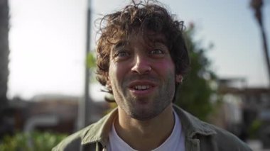 Tüylü saçlı genç bir adam şehir parkında sürpriz ve neşe dolu bir ortam yaratarak doğal ışıklandırma ve yeşil arka planda bir sokak havası yakalıyor..