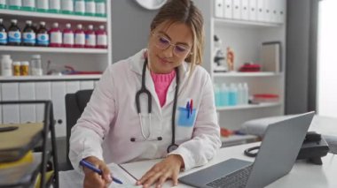 Kadın doktor, hastane ofisindeki masaya dizüstü bilgisayar ve tıbbi malzemelerle notlar yazıyor, evrak işlerine odaklanıyor ve gözlük takıyor, profesyonel sağlık ortamını yansıtıyor..