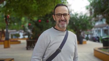 Olgun İspanyol bir adam, arka planda ağaçlar ve binalar olan bir şehir parkında barış işareti yapıyor, yeşillik ve dostça bir ifade sergiliyor..