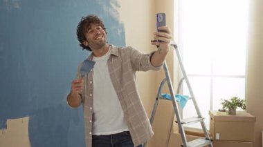 Genç adam resim odasında akıllı telefonuyla selfie çekiyor. Yeni evinde merdivenin yanında duruyor.