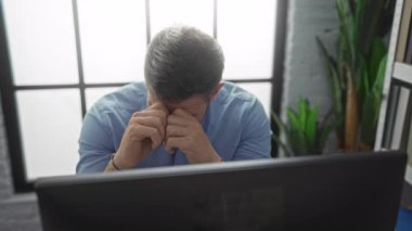 Ofis ortamında çalışan bir adam, ciddi bir ifadeyle, bir bilgisayarın önünde oturmuş, arkasında büyük pencereler ve bitkiler olan modern bir iç mekan sergiliyor..