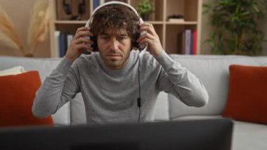 Samimi bir oturma odası ortamında kulaklık takan genç adam, modern iç dekorasyon ve yumuşak yastıklarla çevrili, nötr bir ifadeyle ekrana odaklanmış..