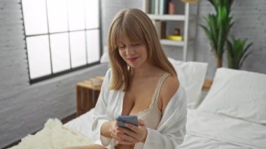 Yatak odasında, rahat bir yatak odasında akıllı telefon kullanan ve doğal ışık gösteren bir kadın. Kapalı mekanda huzurlu bir ortam sergiliyor..