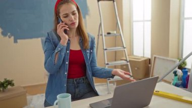 Oturma odasında dizüstü bilgisayar ve yenileme aletleriyle oturan bir kadın telefonda konuşuyor..