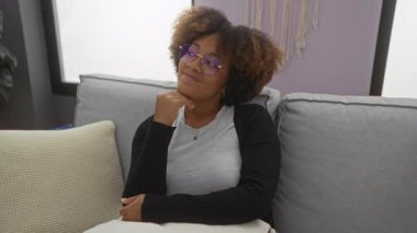 Kapalı alanda gülümseyen gözlüklü ve kıvırcık saçlı bir kadın rahat bir oturma odasında oturuyor..