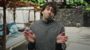 Gri kazaklı genç adam şehir merkezinde yeşil ve kentsel yapıları olan düşünceli bir ifadeyle el çırpıyor..