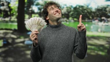 Elinde Rus rublesi tutan İspanyol bir adam parkta neşeli bir ifadeyle gülümsüyor, arka planda doğa filizlenirken başarıyı ve mutluluğu vurguluyor..