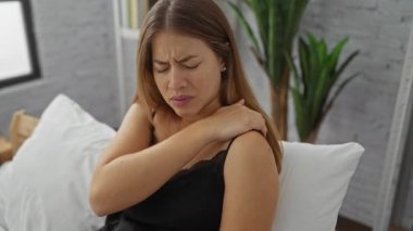 Omuz ağrısı çeken bir kadın yatak odasında rahat bir ortamda beyaz yastıklar ve ev bitkileriyle rahat bir şekilde oturur..