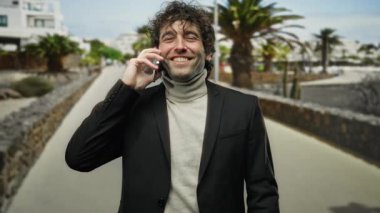 Güneşli bir şehir caddesinde telefonda konuşurken gülümseyen bir adam patikada ağaçlar var, rahat ve kendinden emin bir şehir havası yakalıyor..