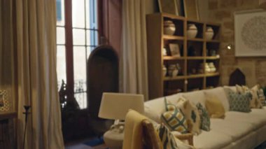 Konforlu dekorlu oturma odası, beyaz kanepe, desenli yastıklar, çömlekli raflar, ve geniş pencereli rahat bir ev ortamı yaratıyor..