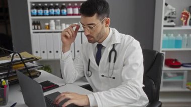 Klinikte gözlük takan genç bir adam laptoptaki tıbbi belgeleri inceliyor. Etrafı şişe ve tıbbi malzemelerle çevrili..