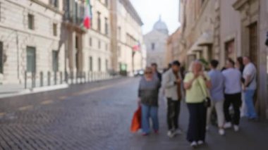 Roma 'da güneşli, bulanık bir kaldırım taşı caddesinde yürüyen insanlar, İtalya' da tarihi binalar ve İtalyan bayrakları ile dolu, hareketli şehir atmosferini ve kültürel zenginliği yakalıyorlar..