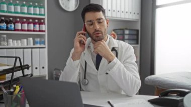 Hastane ofisinde steteskopla konuşan genç bir adam profesyonel çevrede tıbbi malzeme ve dosyalarla çevrili telefonla konuşuyor.