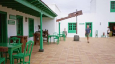 Lanzarote 'deki bulanık koloni tarzı avluda yürüyen bir adam, İspanyol yeşil aksanı, ahşap mobilya ve arka planda bazı insanlar var.