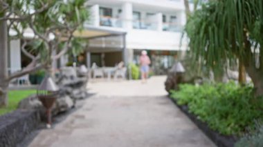 Arka planda yemyeşil ve modern binalar olan tropikal bir otelde yürüyen birinin bulanık görüntüsü..