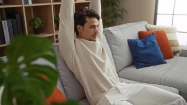 Modern bir oturma odasında rahat bir kanepeye uzanan İspanyol bir adam. Etrafında renkli yastıklar ve bitkiler var. Rahat ve rahat bir ev atmosferi yaratıyor..