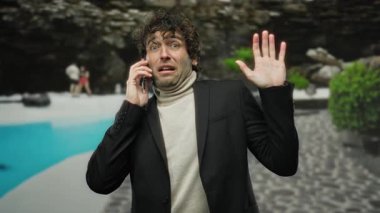 Otelin havuzunun yanında duran, takım elbiseli, dışarıda telefonla konuşurken endişeli görünen, yemyeşil ve rahat arka plan aktiviteleri olan bir adam..
