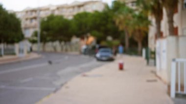 Şehirdeki binalar, ağaçlar, kaldırımlar ve bokeh ışığının altında park edilmiş arabaların olduğu bulanık bir sokak manzarası odağı olmayan bir adamın yürüdüğü rüya gibi bir atmosfer yaratıyor.