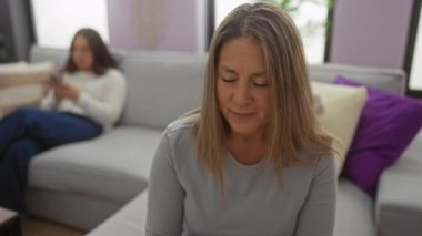 Oturma odasındaki bir kanepede oturan bir kadın, kızı aile dinamiklerini ve iletişim sorunlarını yansıtan bir cep telefonu kullanırken başka tarafa bakıyor..